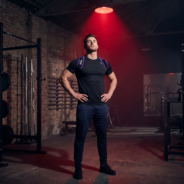 Man wearing PHYSIOATHLETX black and purple posture support brace in an industrial gym setting, upright confident posture